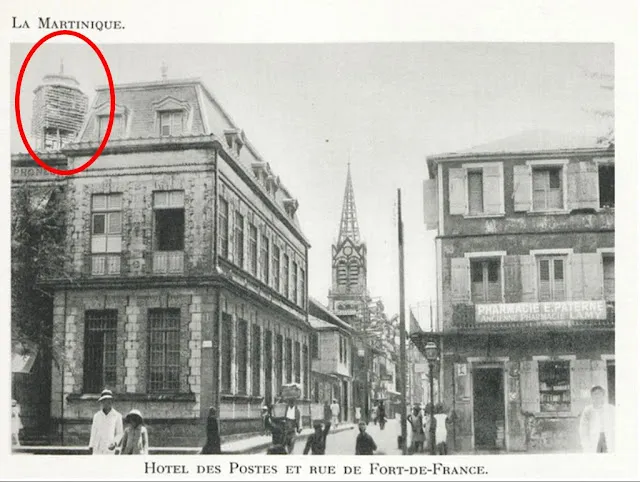 Photographie d'une rue de Fort de France avec des piétons; façade de l'hôtel des postes et d'une Pharmacie avec en arrière plan le clocher de la cathédrale de Fort de France